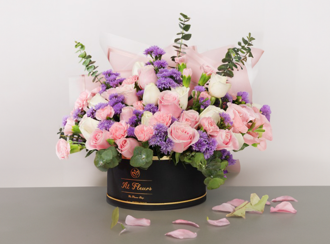 Bouquet of pink and purple flowers in a black box with scattered petals on a table.