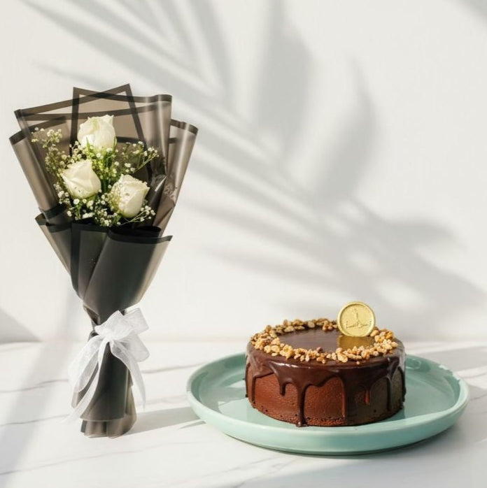 Chocolate cake with a gold coin on a light blue plate next to a bouquet of white flowers on a white surface. Bento Cake and Flower Bouquet