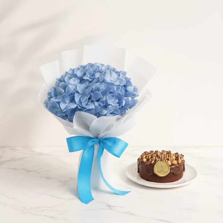Bouquet of blue flowers with a blue ribbon next to a chocolate cake on a white marble surface. Bento Cake and Flower Bouquet