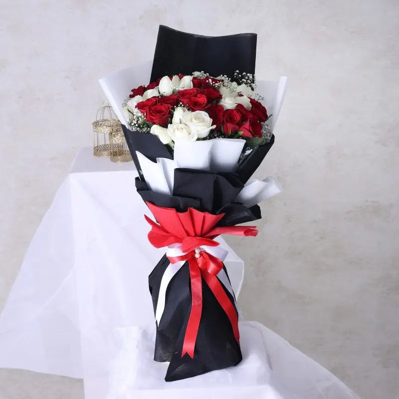A hand bouquet of red and white roses, wrapped in black and red paper with a red ribbon, displayed on a white surface.