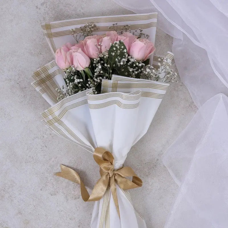A hand bouquet of pink roses wrapped in white and gold striped paper with a ribbon.