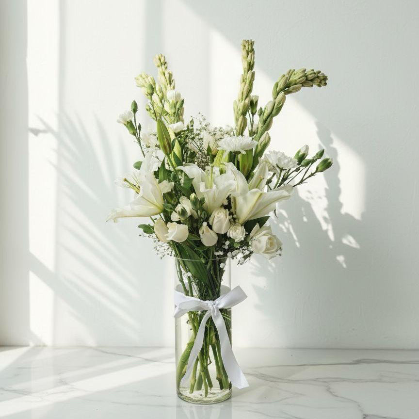 Bouquet of white flowers in a clear vase with a white ribbon on a light surface.