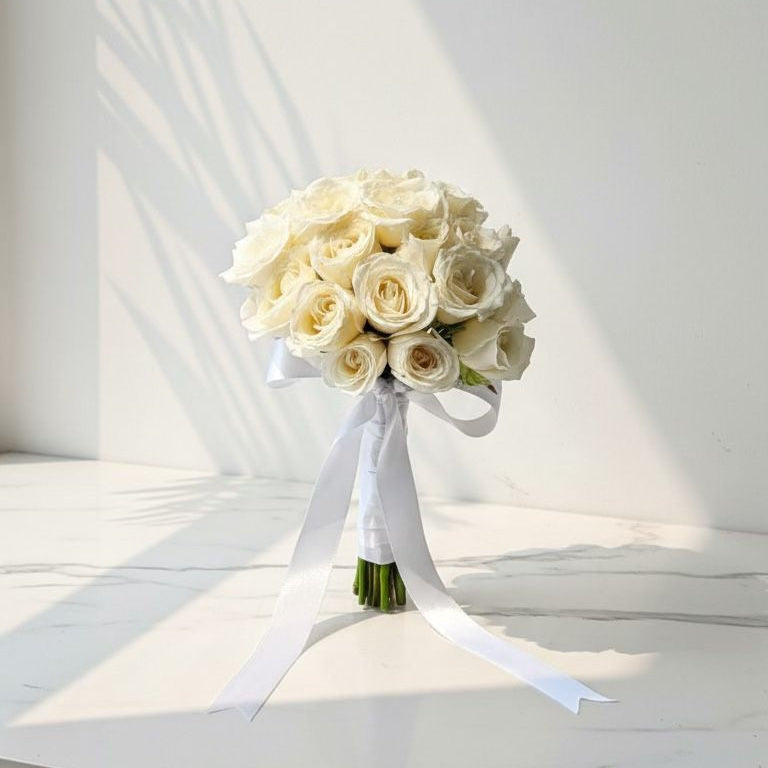 Hand tied white rose bridal bouquet with long white satin ribbon, placed on a marble surface under soft daylight.