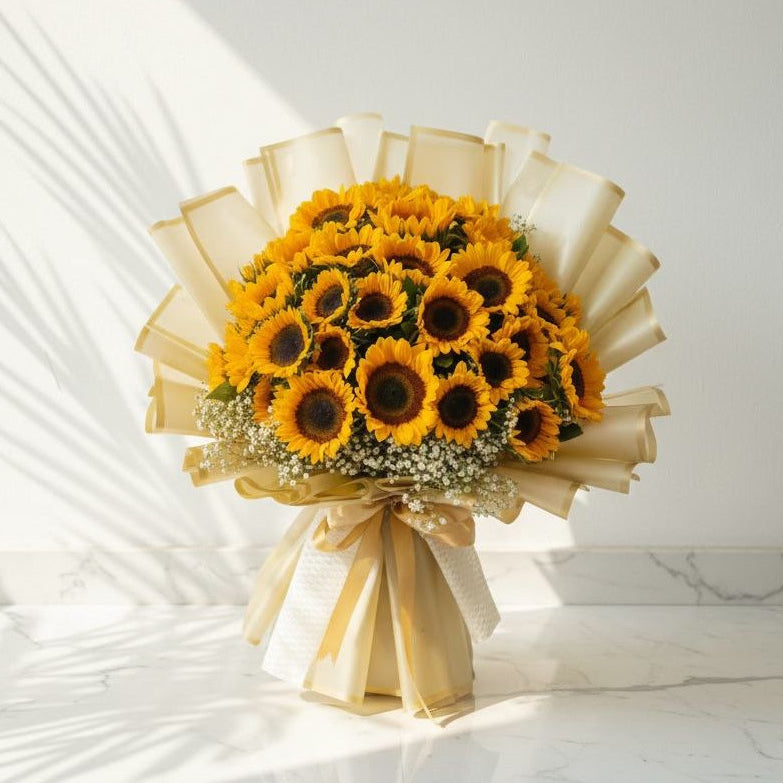 Large bouquet of bright yellow sunflowers accented with white baby’s breath, beautifully wrapped in cream paper with gold detailing and tied with a satin golden ribbon, placed on a marble surface under natural sunlight.