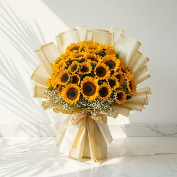 Large bouquet of bright yellow sunflowers accented with white baby’s breath, beautifully wrapped in cream paper with gold detailing and tied with a satin golden ribbon, placed on a marble surface under natural sunlight.