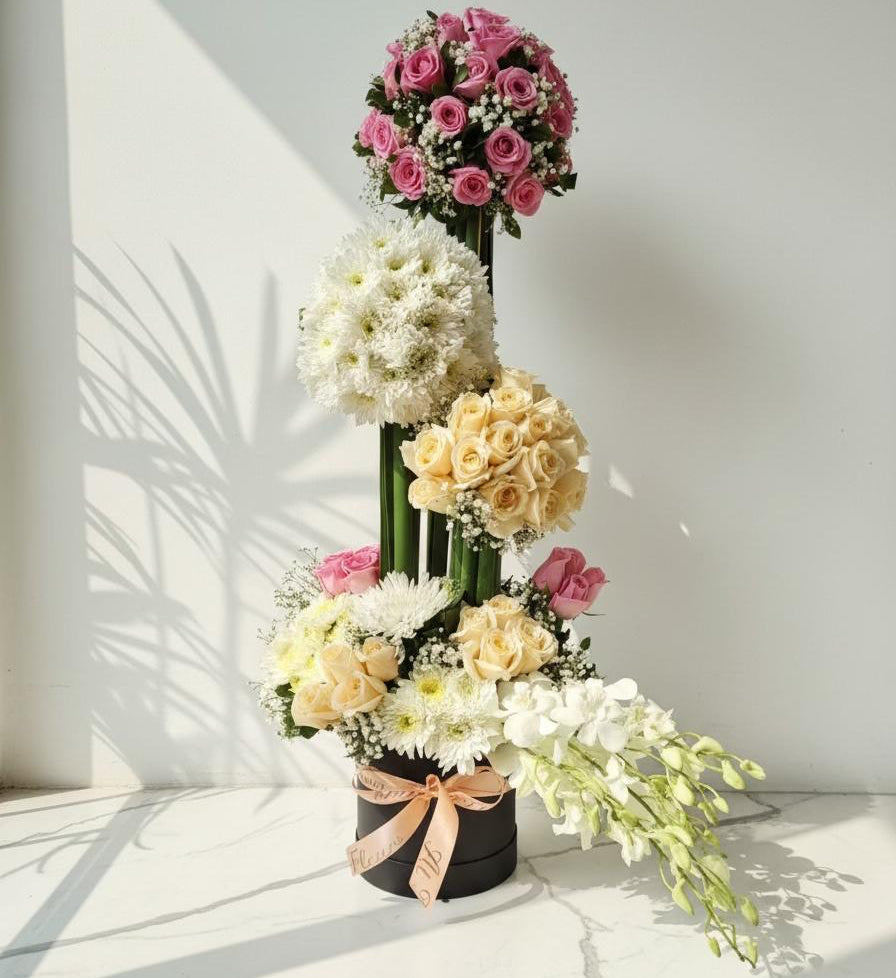 Tall three-layer flower arrangement with pink roses, white chrysanthemums, peach roses, and cascading orchids in a black box.