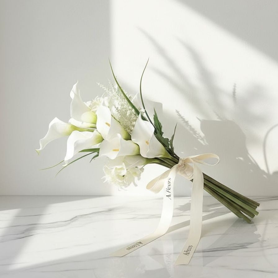 White calla lily bridal bouquet with long stems, tied with an ivory satin ribbon that has “AtFleurs” printed on it, placed on a marble surface under soft sunlight.