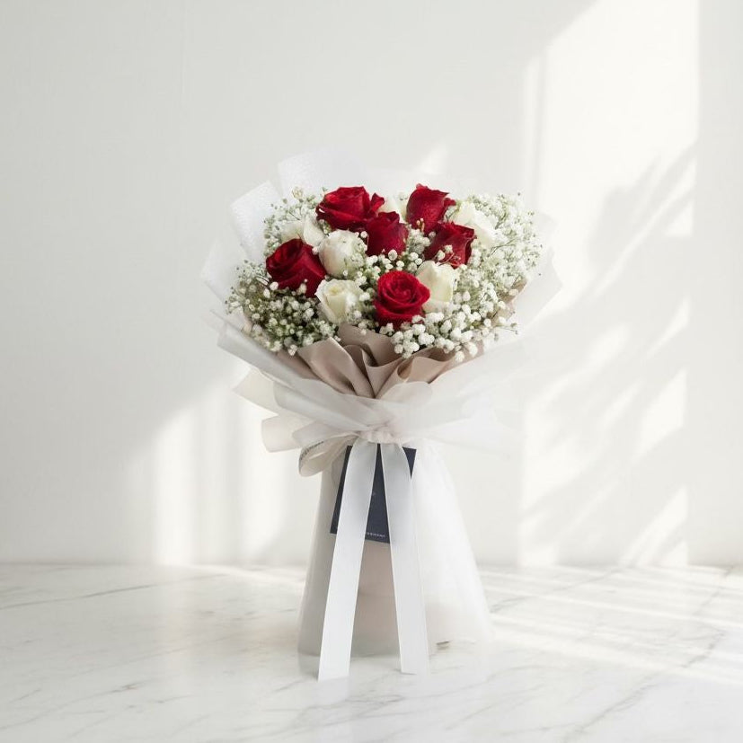 Bouquet of red and white roses with baby’s breath, wrapped in white and beige paper and tied with a long white satin ribbon, placed on a marble surface under sunlight.