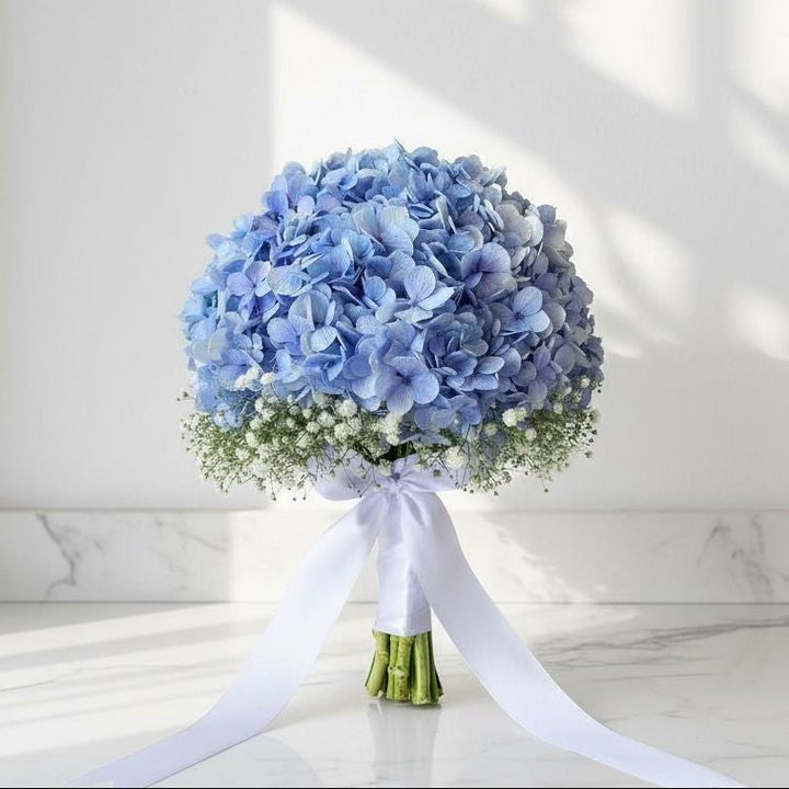 Round hand bouquet of blue hydrangeas surrounded by white baby’s breath, tied with a long white satin ribbon, placed on a marble surface under natural light.