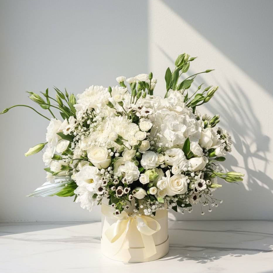 Round box arrangement with fresh white roses, carnations, chrysanthemums, lisianthus, hydrangeas, and baby’s breath, tied with a cream ribbon.