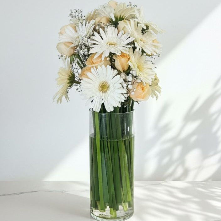 White gerberas, peach roses, and baby’s breath arranged in a tall glass vase with green stems visible, placed on a marble surface under natural light.