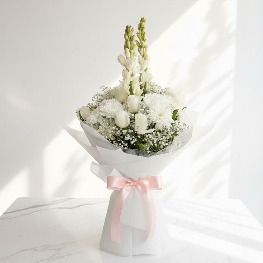 White floral bouquet with roses, tuberose, and chrysanthemums, wrapped in white paper and tied with a soft pink satin ribbon, placed on a marble surface under natural light.