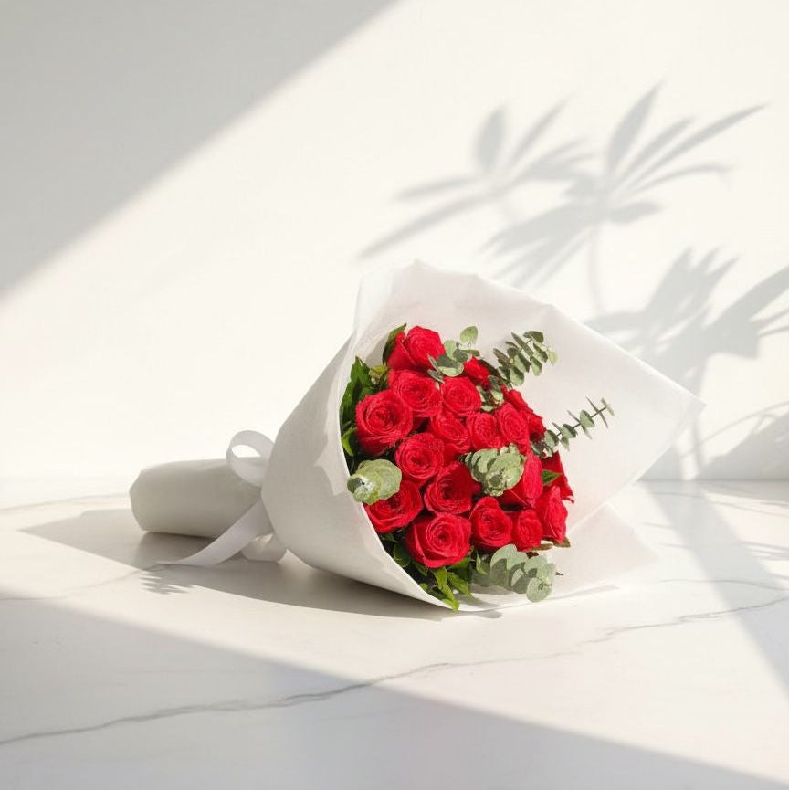 Bouquet of fresh red roses with eucalyptus leaves, wrapped in white paper and tied with a ribbon, placed on a marble surface under natural sunlight