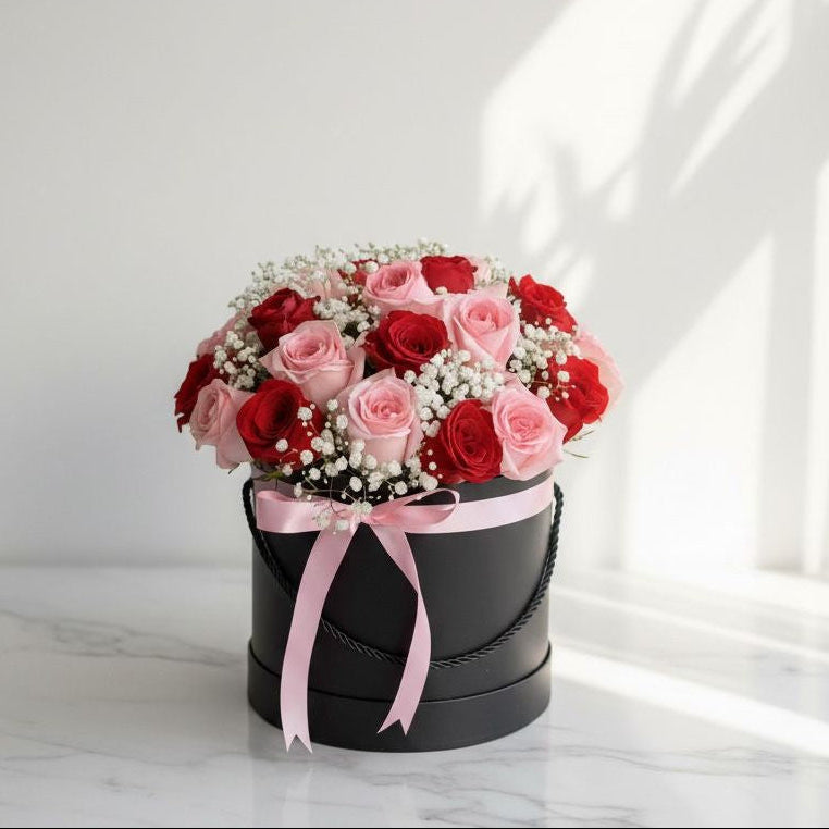 Round black flower box filled with red and pink roses accented with white baby’s breath, finished with a light pink satin ribbon, placed on a marble surface under natural sunlight.