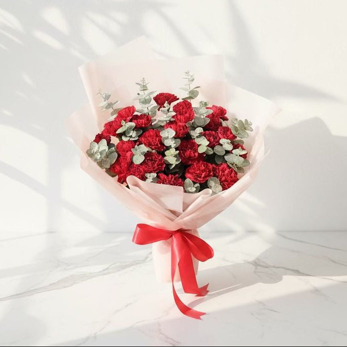 Bouquet of red carnations with eucalyptus leaves, wrapped in soft pink paper and tied with a red satin ribbon, placed on a marble surface under sunlight.
