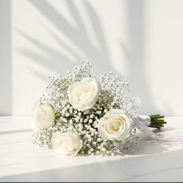 White rose hand bouquet with baby’s breath, tied with a white satin ribbon, placed on a marble surface under natural light