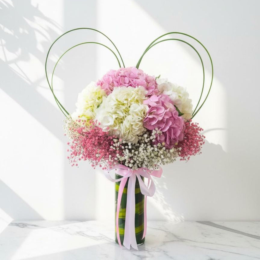 Vase arrangement with pink and white hydrangeas, pink baby’s breath, white baby’s breath, and looped green leaves, tied with a pink satin ribbon.