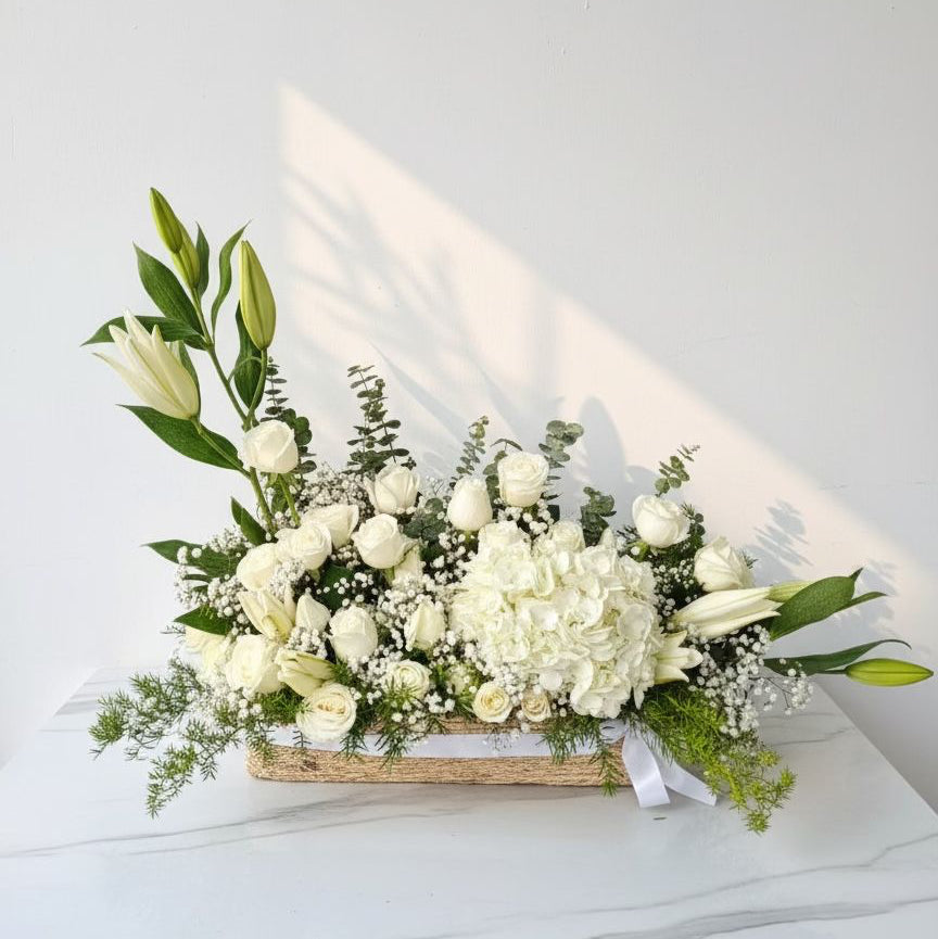 Tall vase arrangement with white lily buds, purple button flowers, eucalyptus, baby’s breath, and greens, tied with a green ribbon.