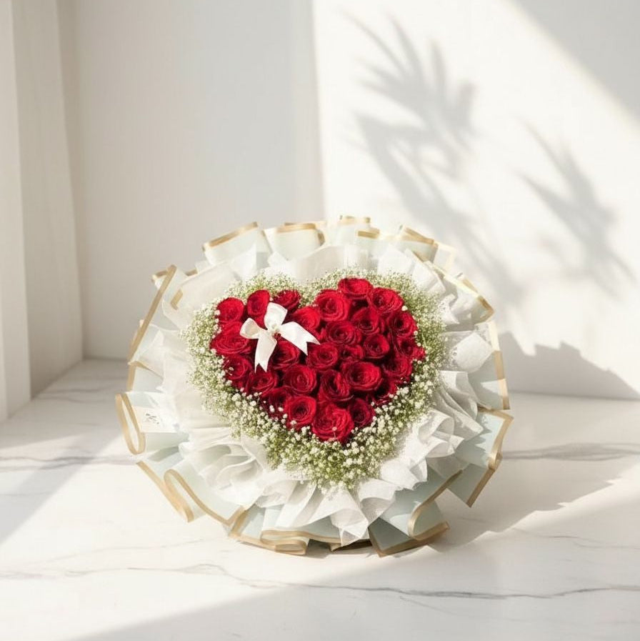 eart-shaped bouquet made with red roses surrounded by white baby’s breath, wrapped in white and gold-edged paper with a small white ribbon bow, placed on a marble surface under sunlight.