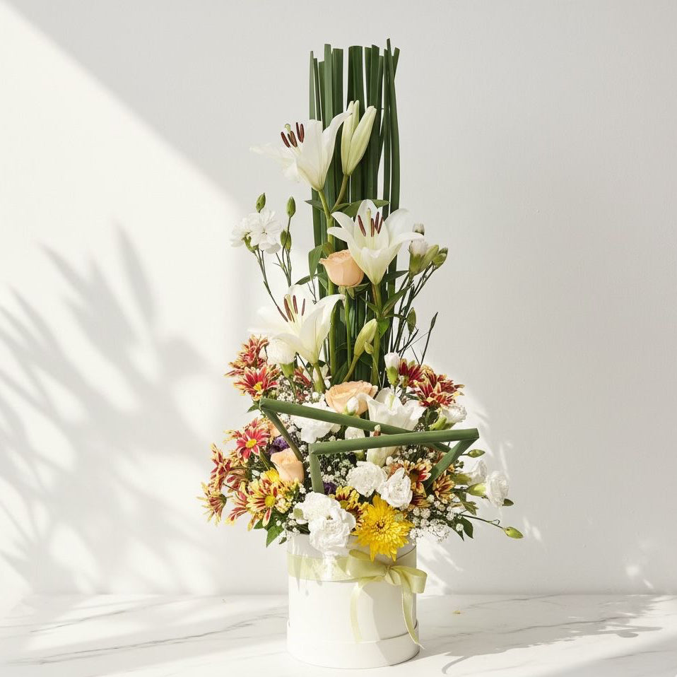 Tall mixed flower arrangement with lilies, carnations, chrysanthemums, and peach roses in a white box.