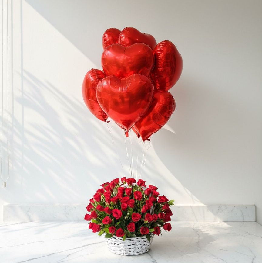 Red heart-shaped balloons and a basket of red roses on a white marble surface with a white wall background.