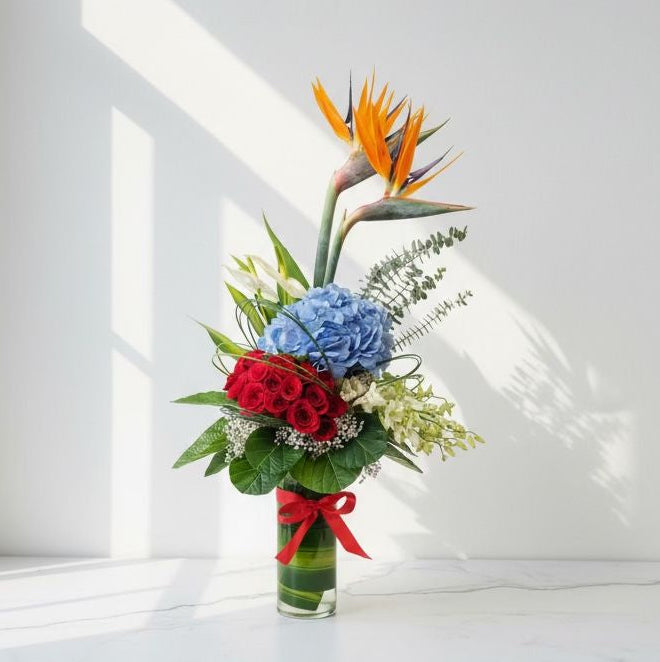 Bouquet of flowers with red roses, blue hydrangeas, and a bird of paradise in a clear vase on a white background.