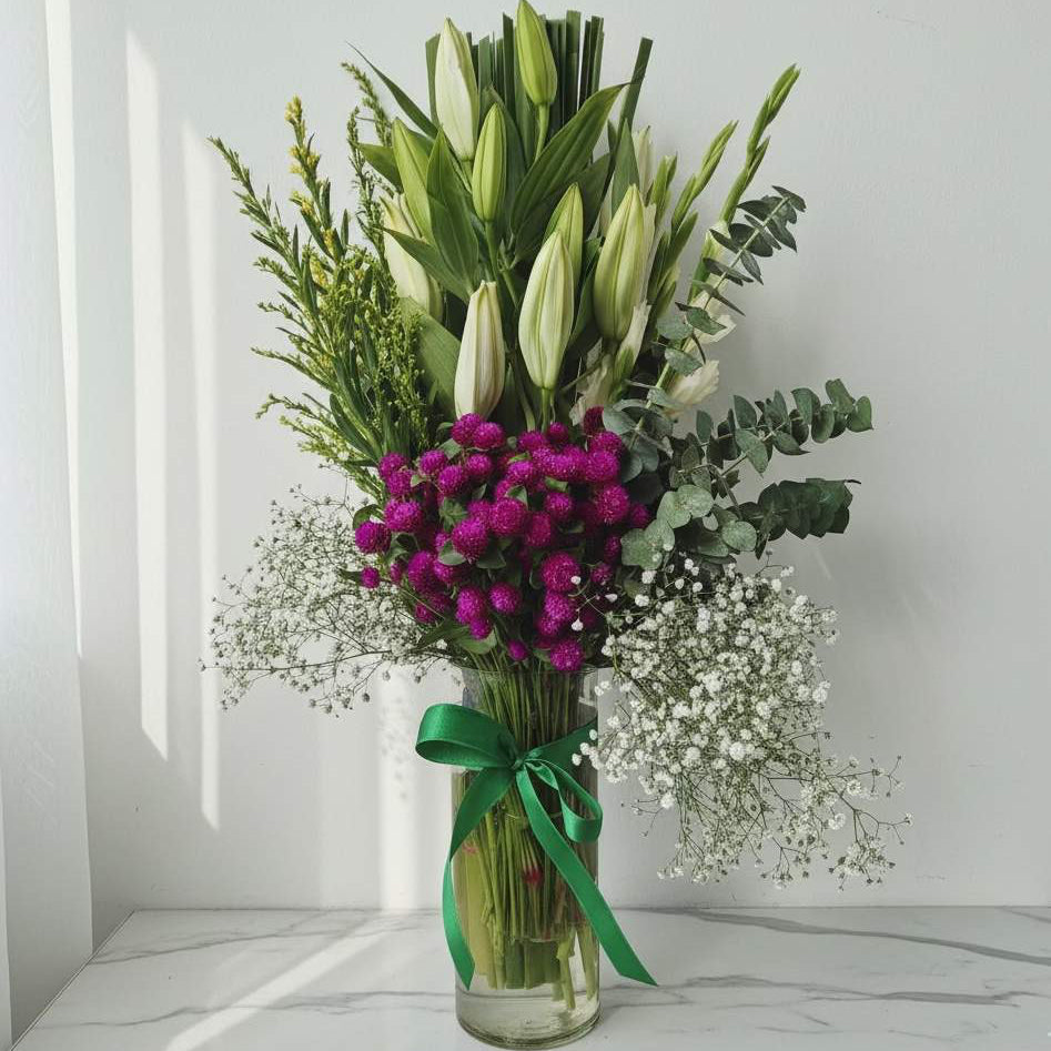 Tall vase arrangement with white lily buds, purple button flowers, eucalyptus, baby’s breath, and greens, tied with a green ribbon.