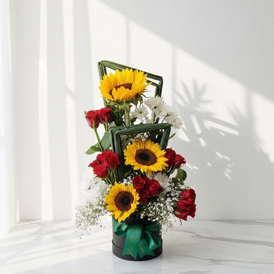 Tall floral arrangement with sunflowers, red roses, white chrysanthemums, baby’s breath, and styled leaves in a black box with a green ribbon.